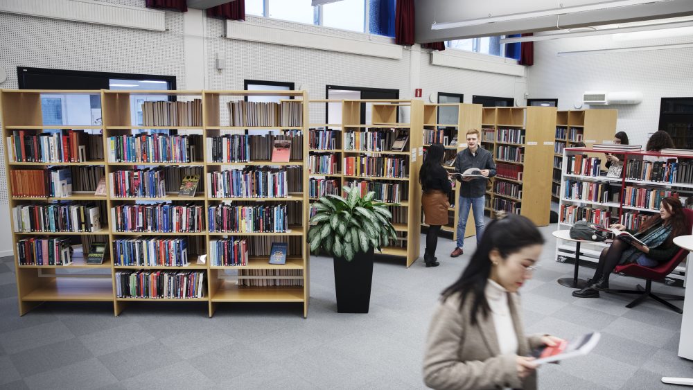 Bookshelves in a library, a woman walking by a book, a couple chatting in the background each holding books.